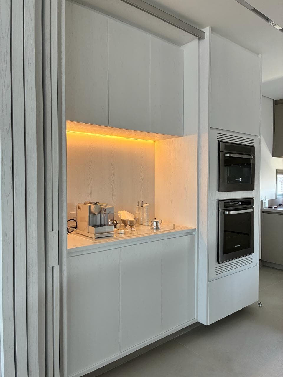 A white kitchen with a coffee station set into a warmly backlit niche, handleless fronts and a built-in double oven column.
