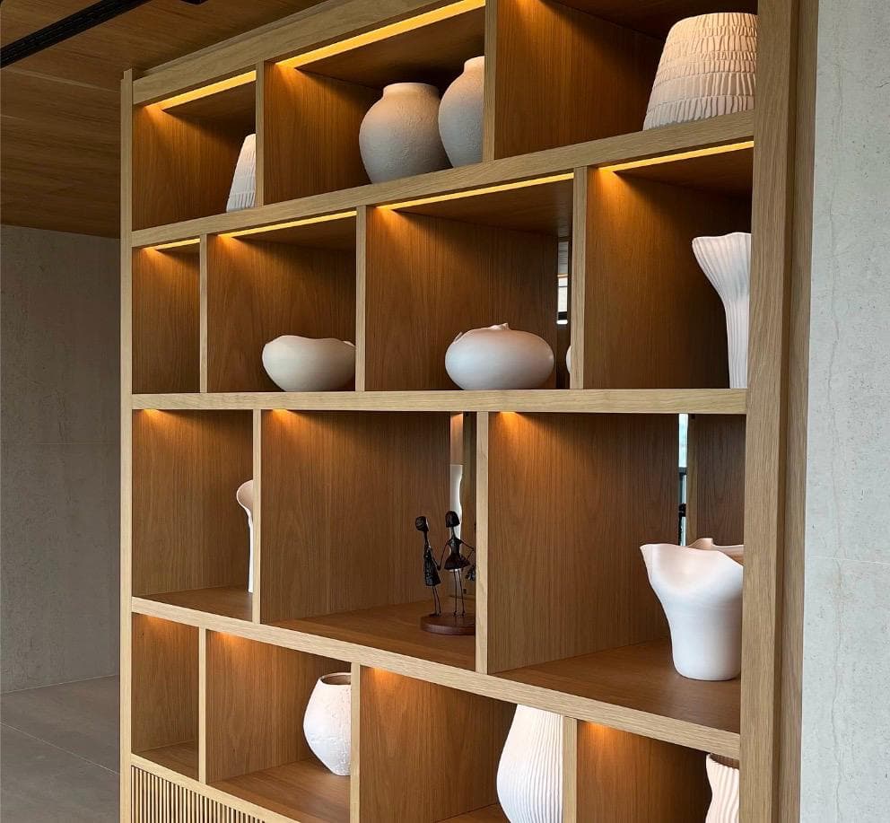An oak open shelving unit used as a room divider, each compartment lit from above and holding pale ceramic vessels.