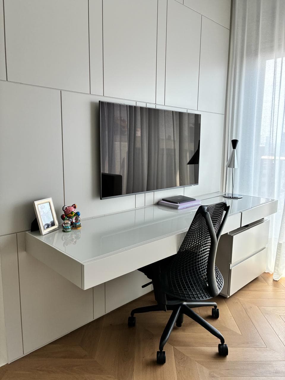 A white lacquered wall of cabinets with a cantilevered glass-topped desk and a wall-mounted screen.