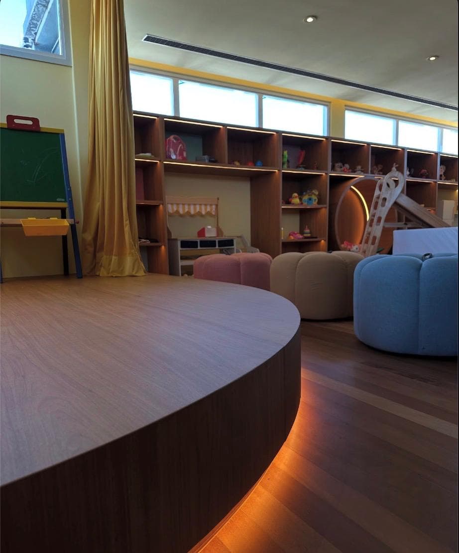 A children's playroom with a curved oak platform lit from beneath and a long wall of lit cubby shelving.